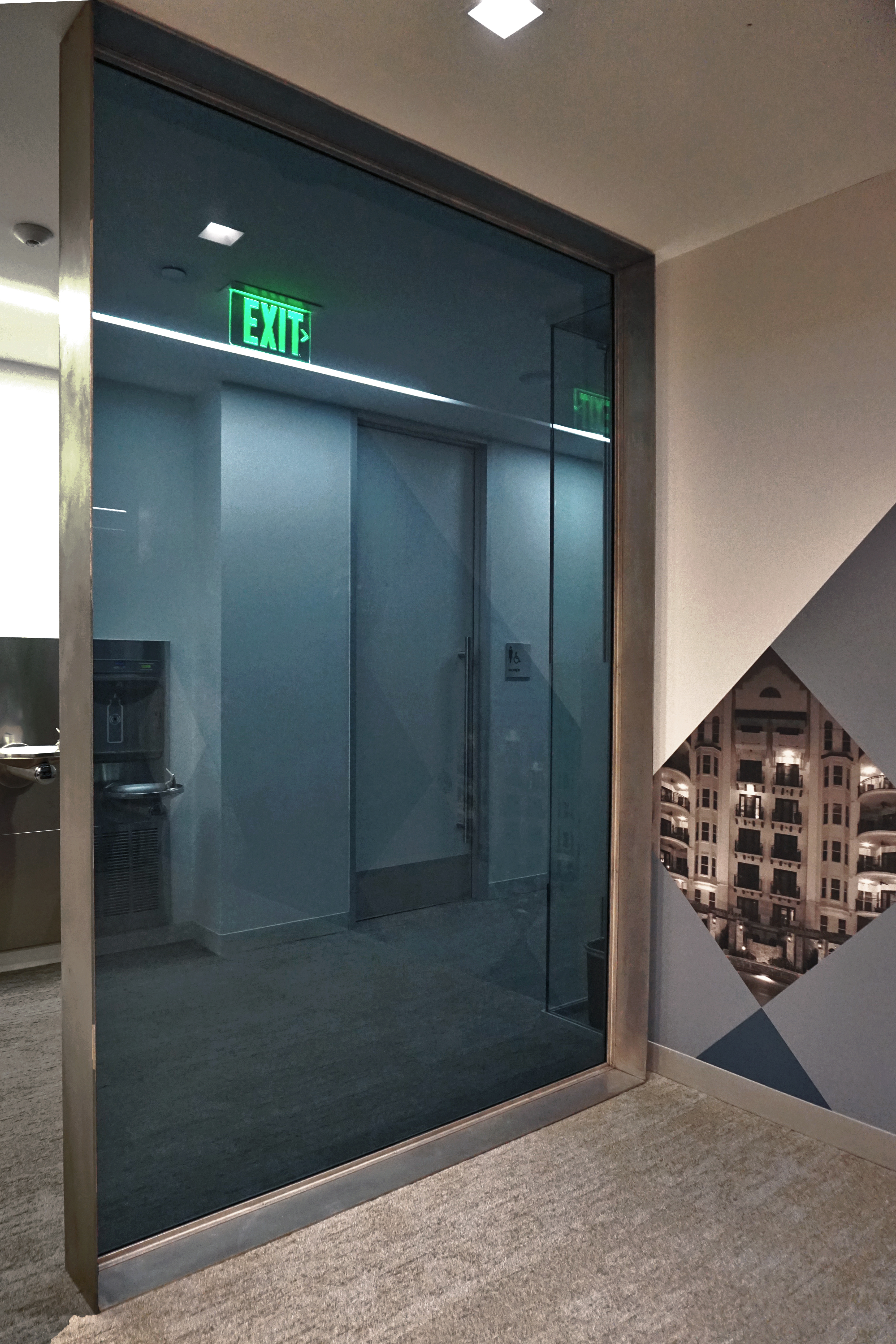 Glass elevator doors with a reflection of the exit sign and interior walls.